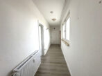 Narrow white hallway with textured walls, light wood floor, radiator on the left, window with a sill on the right, and a door at the far end.