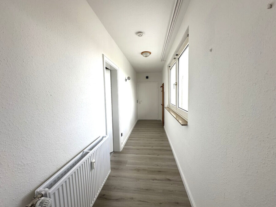 Narrow white hallway with textured walls, light wood floor, radiator on the left, window with a sill on the right, and a door at the far end.