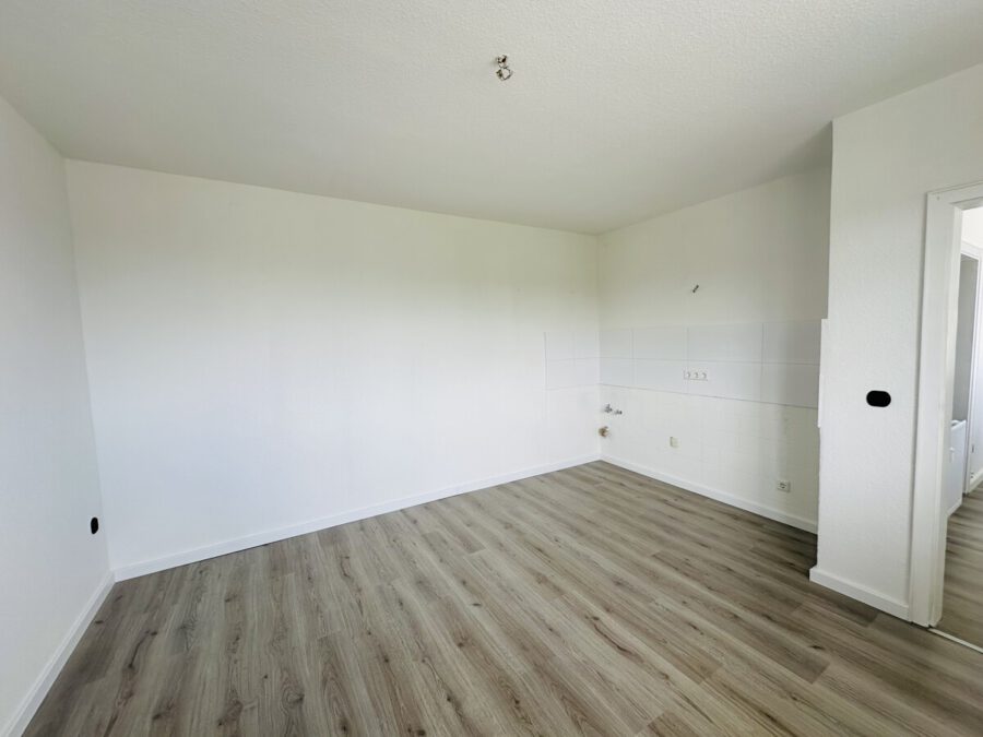 Empty, unfurnished kitchen space with light wood flooring and a white tiled wall area for future cabinets; exposed plumbing near the corner.