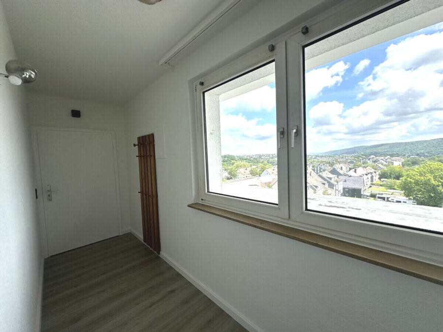Narrow white hallway with a large window showing blue sky and a town below; door at the far end and a wooden folding screen leaning against the wall.