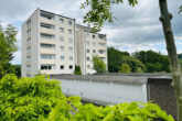 White multi-story apartment building with balconies, surrounded by trees and shrubs, under a cloudy sky.