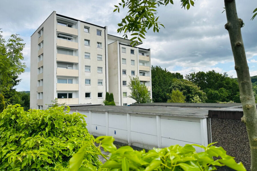 White multi-story apartment building with balconies, surrounded by trees and shrubs, under a cloudy sky.