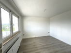Empty, unfurnished room with white walls, wide windows along one wall, a radiator beneath the window, and a light wooden floor; ceiling has exposed wires for a future fixture.