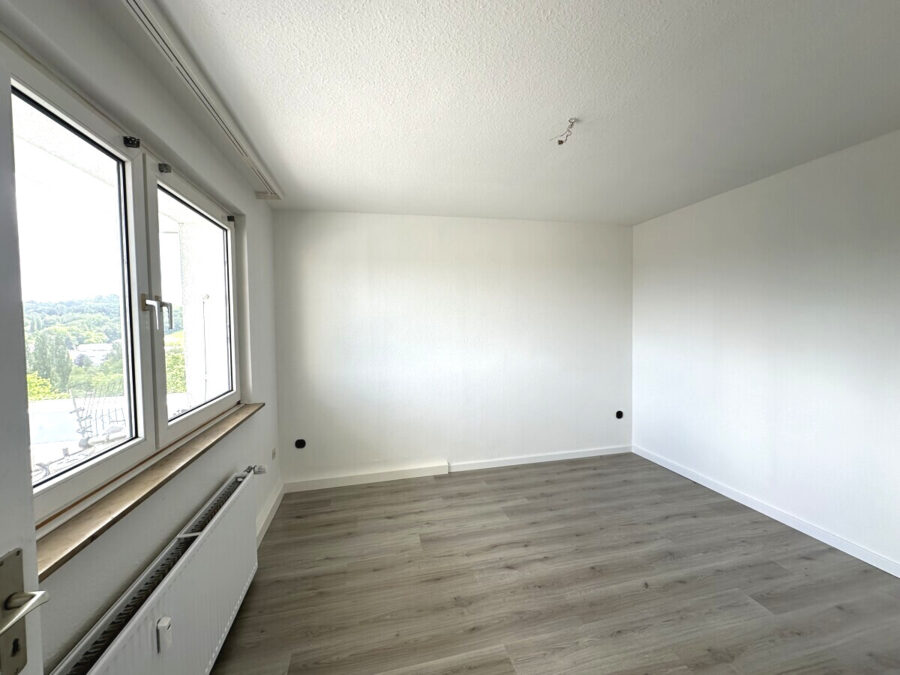 Empty, unfurnished room with white walls, wide windows along one wall, a radiator beneath the window, and a light wooden floor; ceiling has exposed wires for a future fixture.