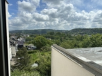 View from a balcony overlooking a green town with houses, trees, and rolling hills under a cloudy sky.