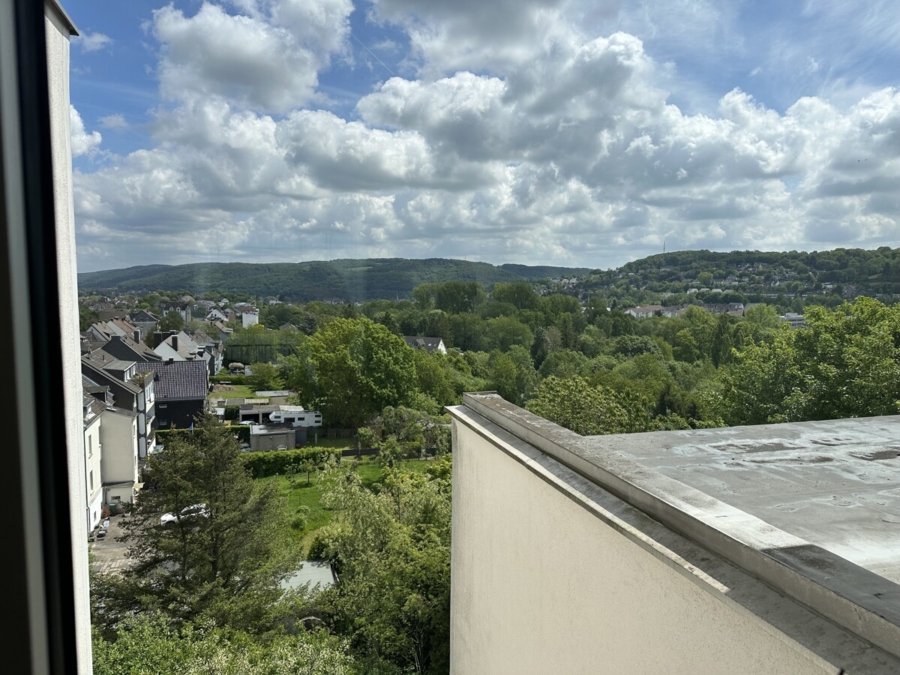 View from a balcony overlooking a green town with houses, trees, and rolling hills under a cloudy sky.
