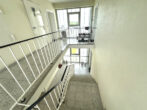 Interior stairwell landing with white walls, metal railings, and tiled floors; doors line the corridor to the left and a window at the far end floods the space with light.