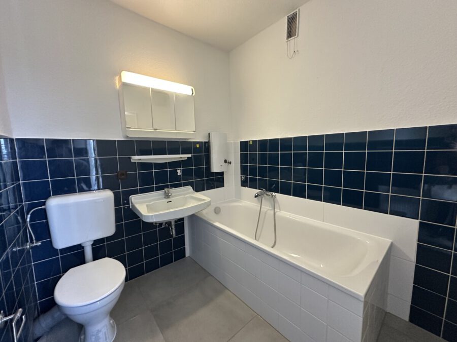 White bathtub with chrome fixtures against dark blue tiled wall, next to a white sink and toilet in a small bathroom