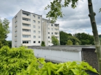 Four-story apartment building with beige balconies and white facade, surrounded by green trees and shrubs under a cloudy sky.