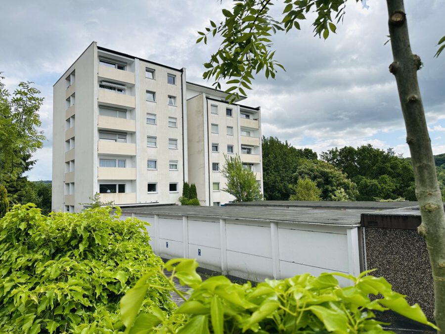 Four-story apartment building with beige balconies and white facade, surrounded by green trees and shrubs under a cloudy sky.